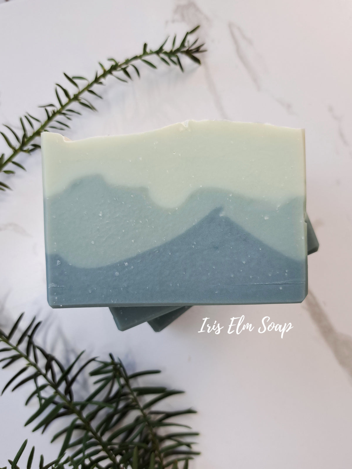 A rectangular bar of layered balsam fir soap with a mountain-like design in pale sage green on top and slate blue on the bottom, styled on a white marble surface with pine branches. The text 'Iris Elm Soap' is visible on the right side.