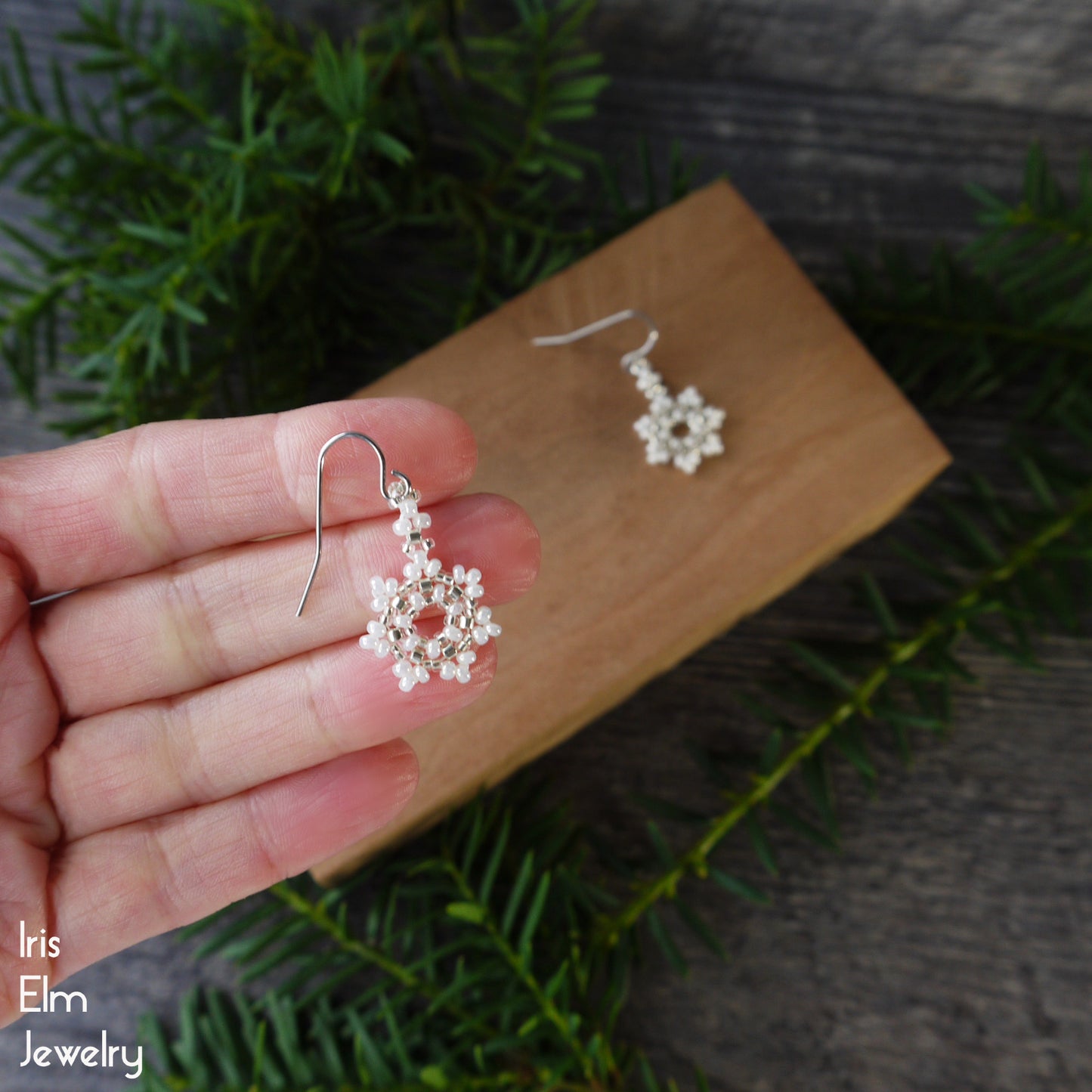 Silver and pearl white glass beaded snowflake earrings held in a hand with a wooden board and greenery in the background, featuring 'Iris Elm Jewelry' branding.