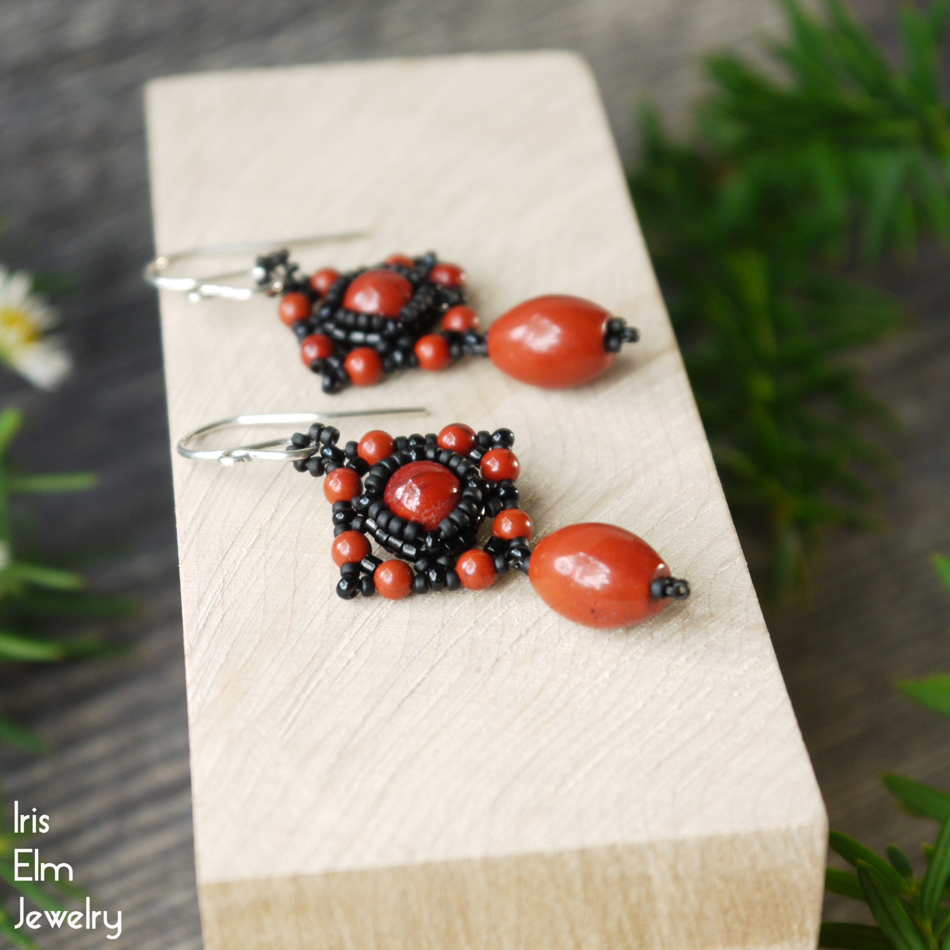 Pair of red jasper and black seed bead beaded earrings on a wooden block with 'Iris Elm Jewelry' branding.