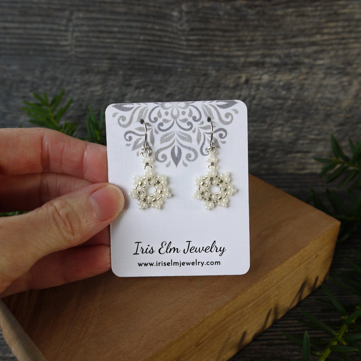 Pair of white snowflake earrings on a 'Iris Elm Jewelry' card held by a hand with a natural background.