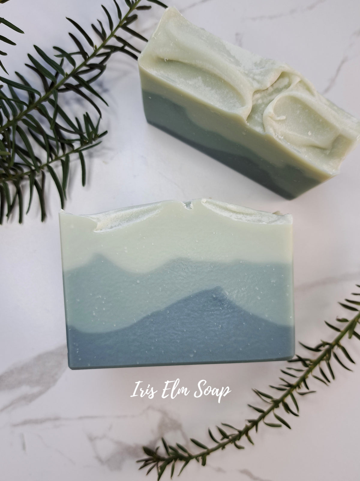 Two rectangular balsam fir handmade soap bars with a layered wave design in pale sage green and soft blue, arranged on a white marble surface with pine branches. The text 'Iris Elm Soap' is visible at the bottom.