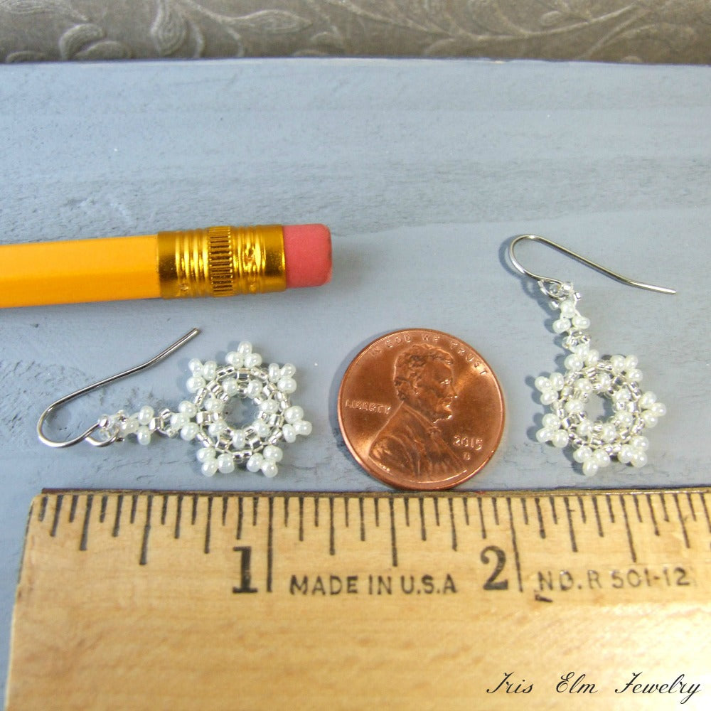 Pair of snowflake earrings with white beads next to a penny and ruler for size comparison on a light blue background.