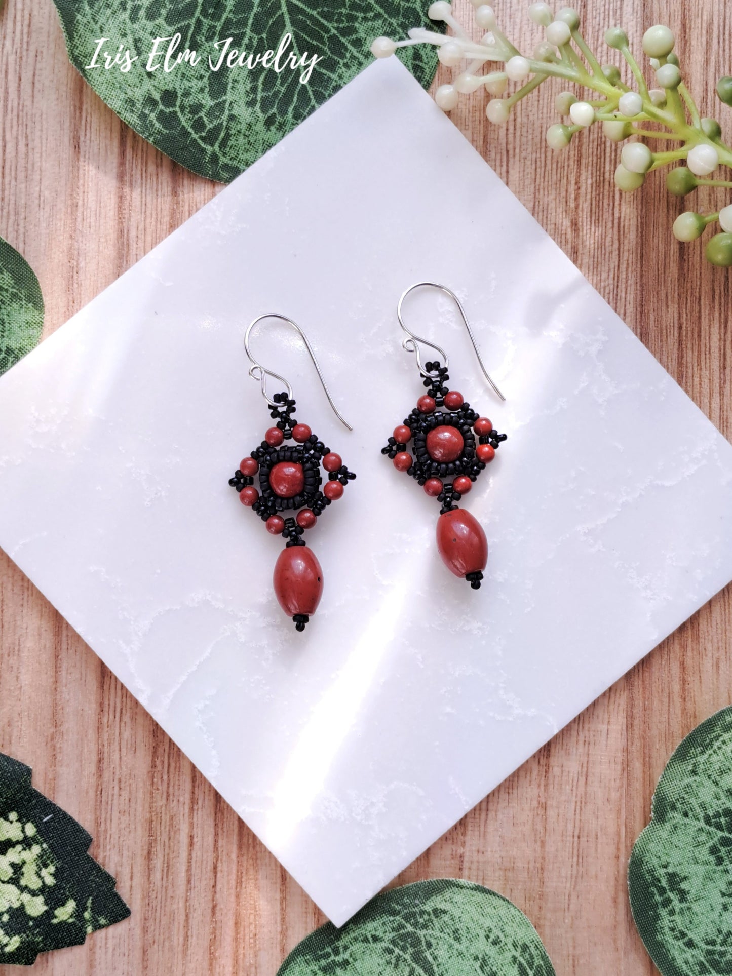 Pair of black and red jasper beaded earrings on a white cloth with green leaves in the background