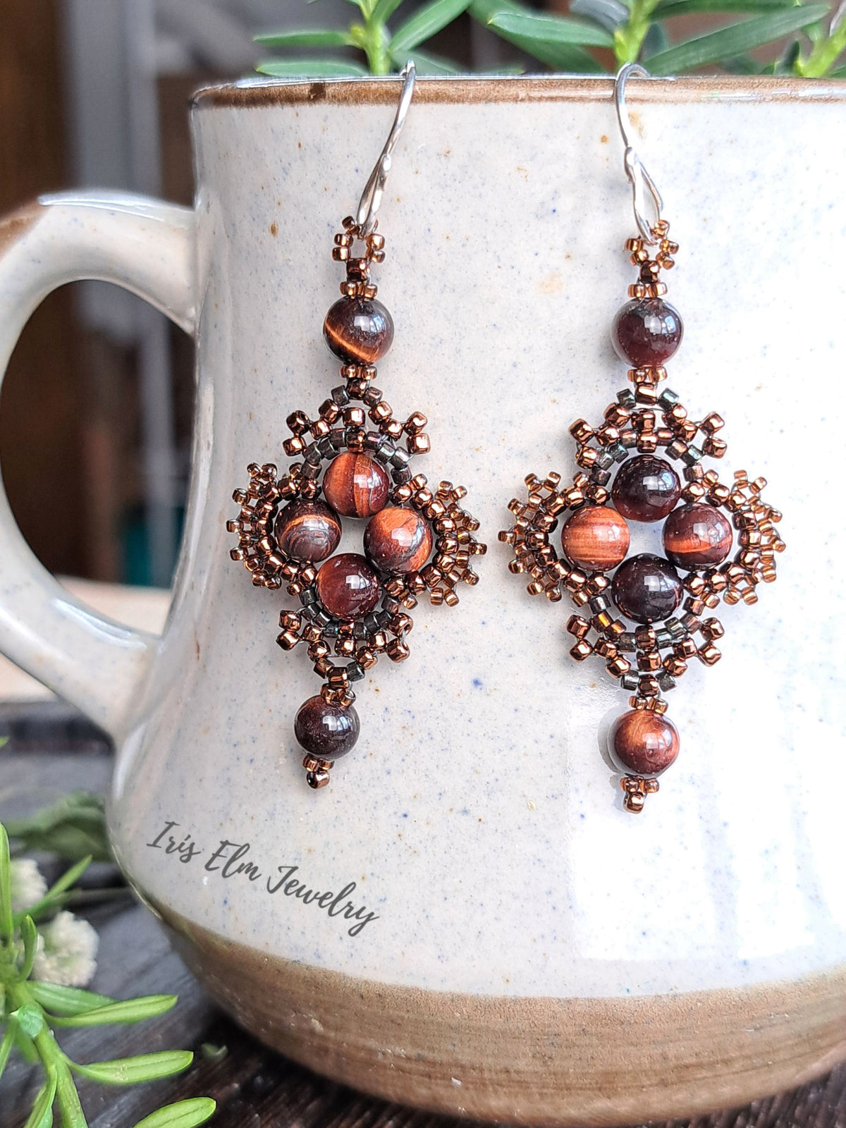 A pair of handmade beaded drop earrings with brown-toned seed bead filigree, red tiger's eye beads, and dark brown beads, hanging in front of a ceramic mug. The brand name 'Iris Elm Jewelry' is printed on the mug.