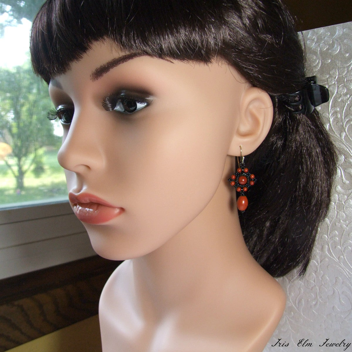 Mannequin head with dark hair styled in a ponytail, wearing red jasper and black earrings.