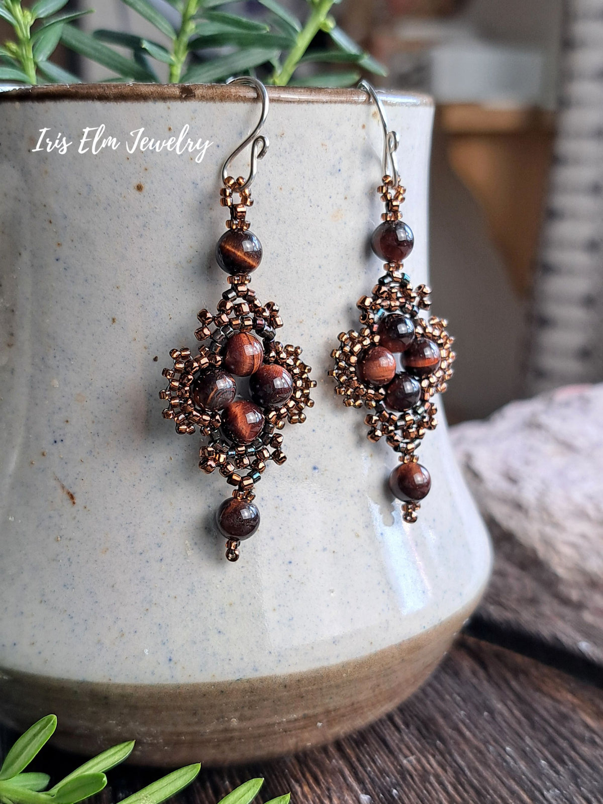A pair of handmade beaded dangle earrings with mahogany red tiger's eye beads and bronze-toned seed beads, hanging in front of a ceramic pot. The brand name 'Iris Elm Jewelry' is visible as a watermark on the left.