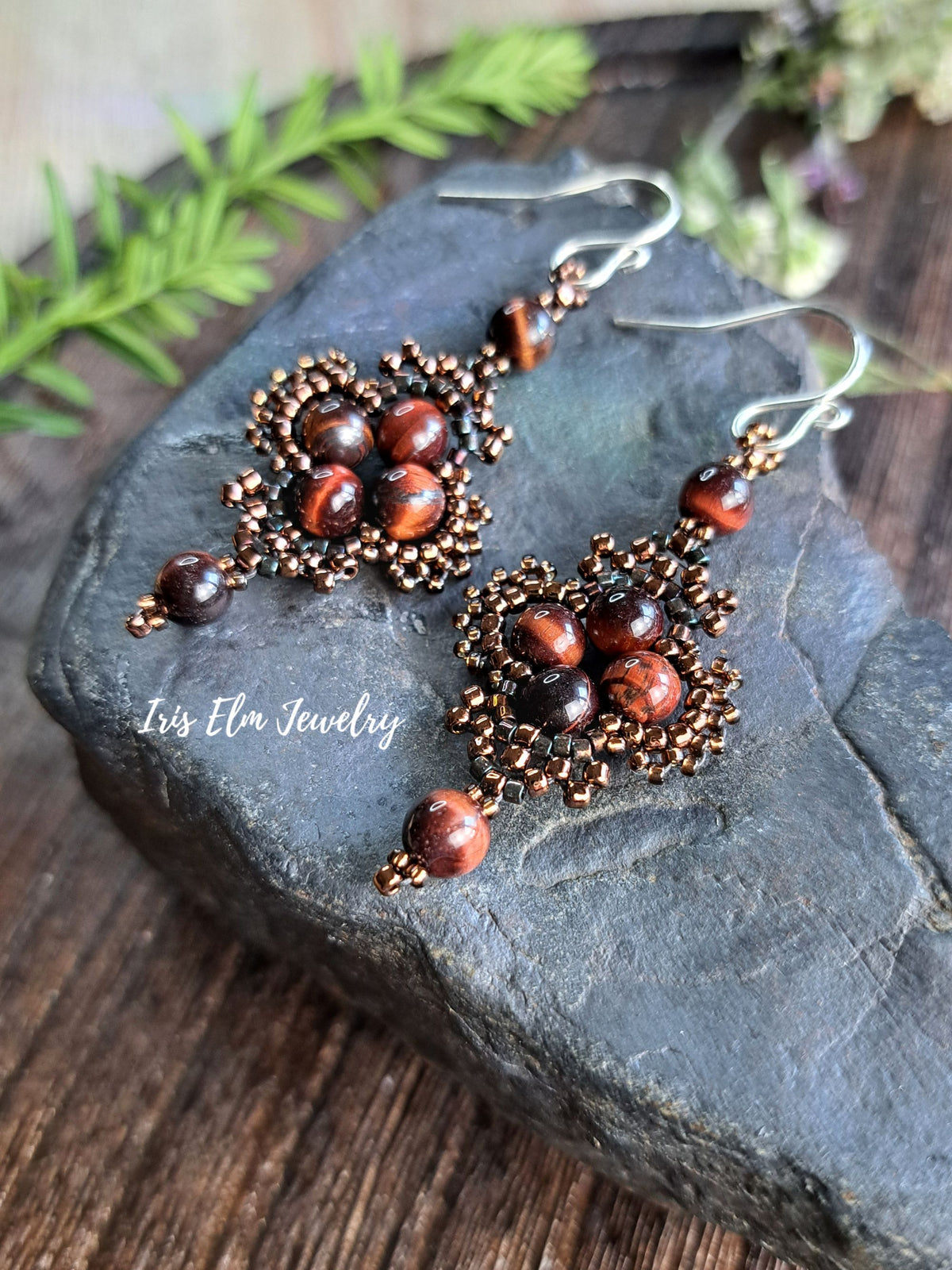 A pair of handmade dangle earrings with brown-toned seed beads and tiger's eye beads, displayed on a dark slate stone with a wooden surface and green foliage in the background. The brand name 'Iris Elm Jewelry' is watermarked on the left.
