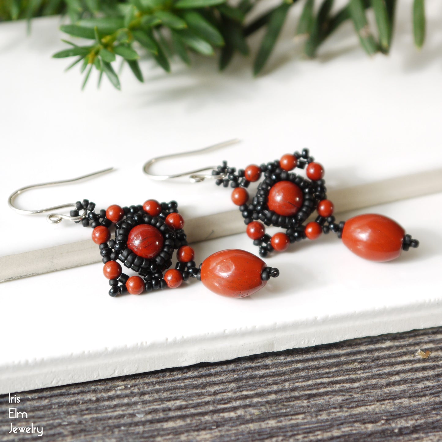 Beaded earrings with red jasper gemstone and black glass beads on a white surface with greenery in the background.