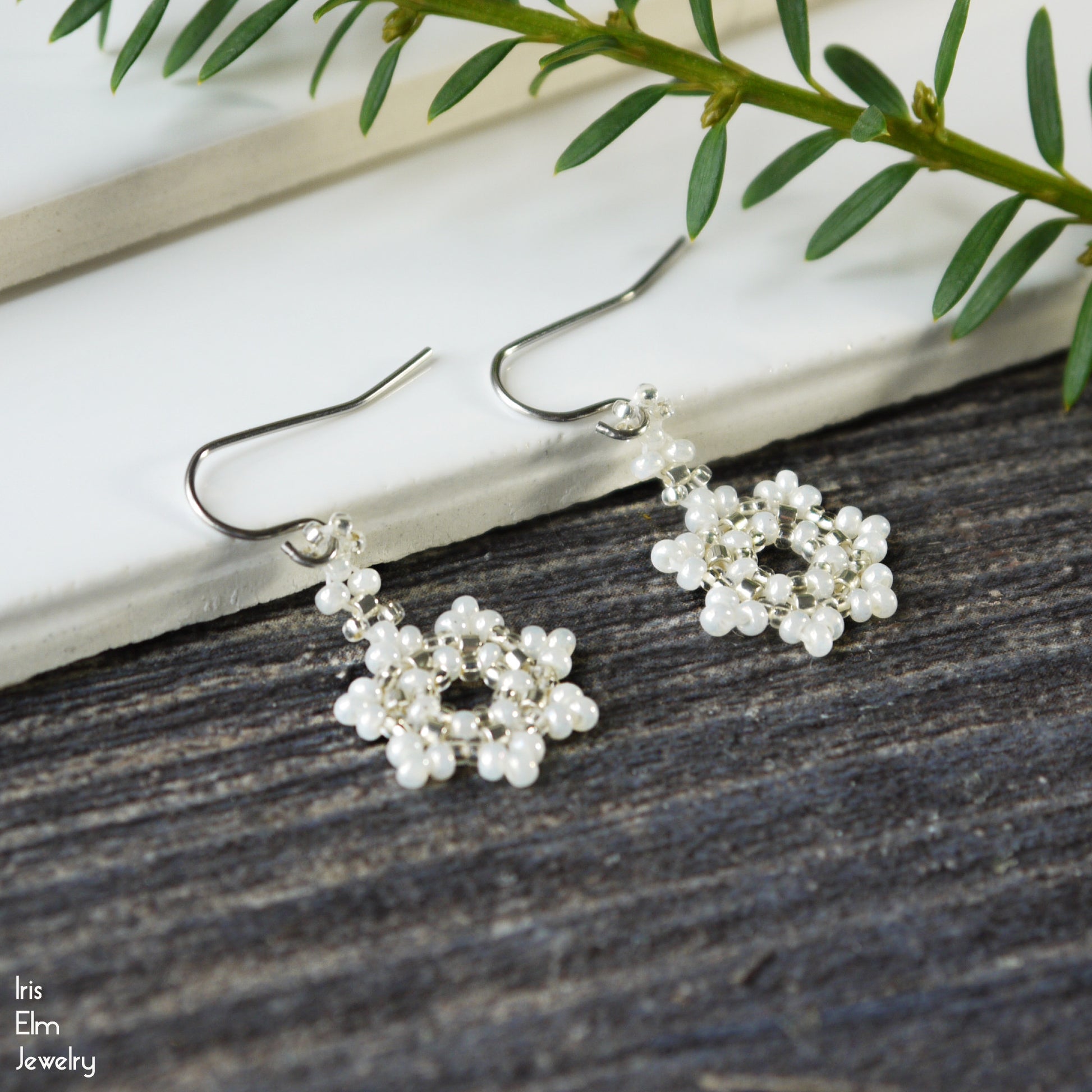 winter snowflake woven seed bead earrings with pearl-like beads on a wooden surface with green leaves.