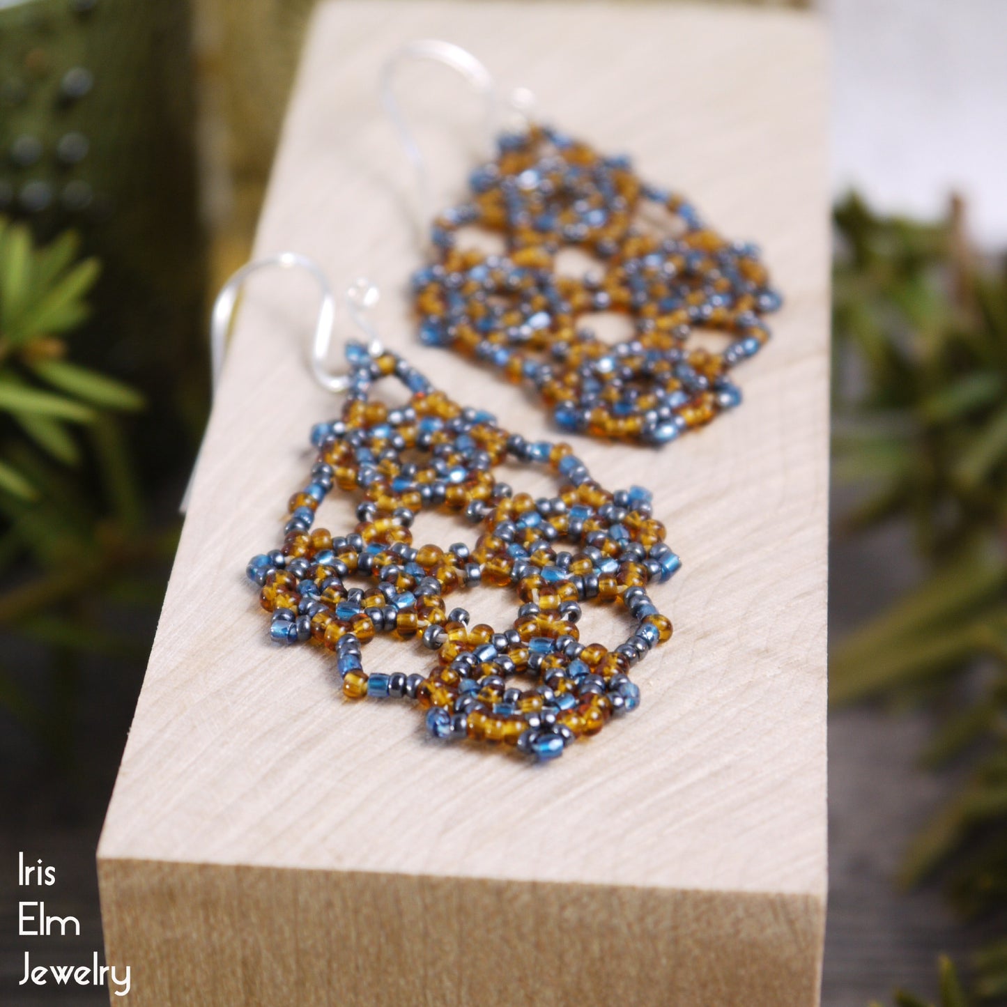 Pair of beaded amber orange and blue seed bead lattice earrings on a wooden block with 'Iris Elm Jewelry' branding.