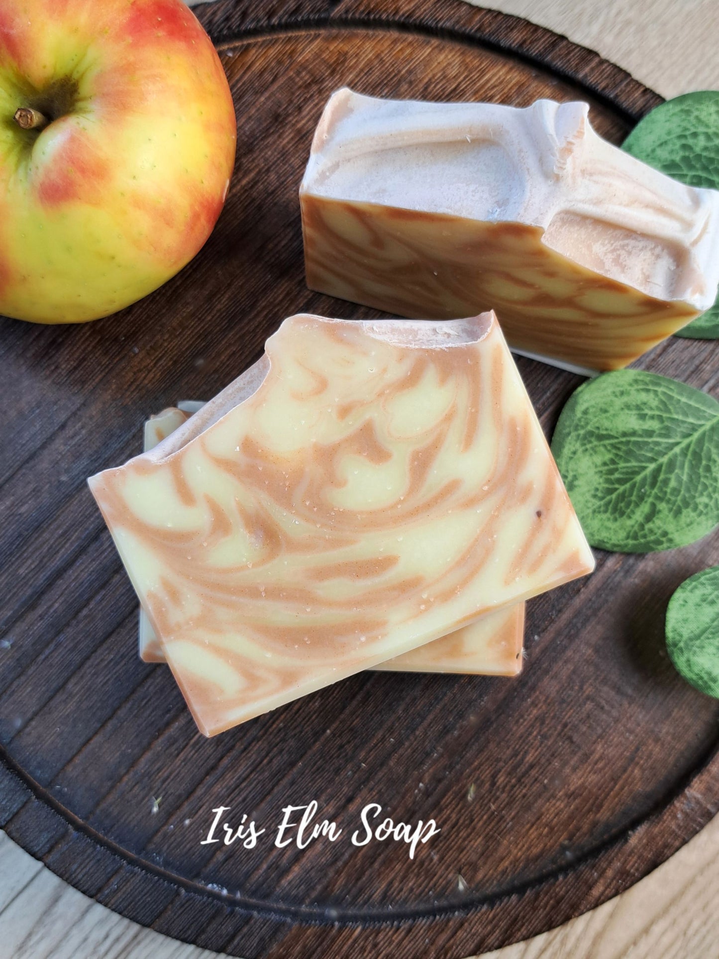 Two apple scented decorative bars of Iris Elm Soap with a marbled cream and light brown swirl pattern, displayed on a dark wooden board next to a red and yellow apple and green leaves.