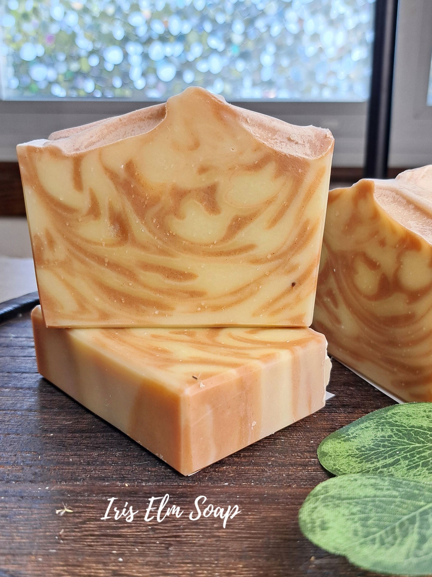 Three handmade apple soap bars with a marbled swirl pattern of cream, beige, and light brown, stacked on a dark wooden surface with a green leaf in the foreground and a window in the background. The text 'Iris Elm Soap' is visible at the bottom.