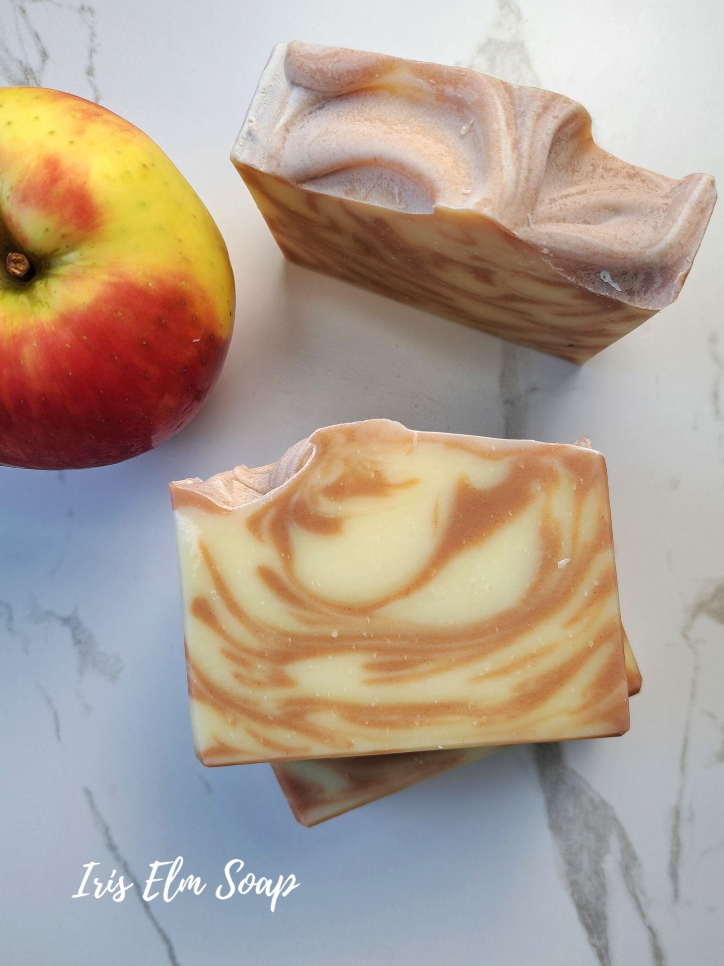 Two rectangular bars of swirled apple scented handmade soap with a creamy off-white and light brown marbled pattern, displayed on a light marble surface next to a red and yellow apple. The brand name 'Iris Elm Soap' is written in white script in the bottom-left corner.
