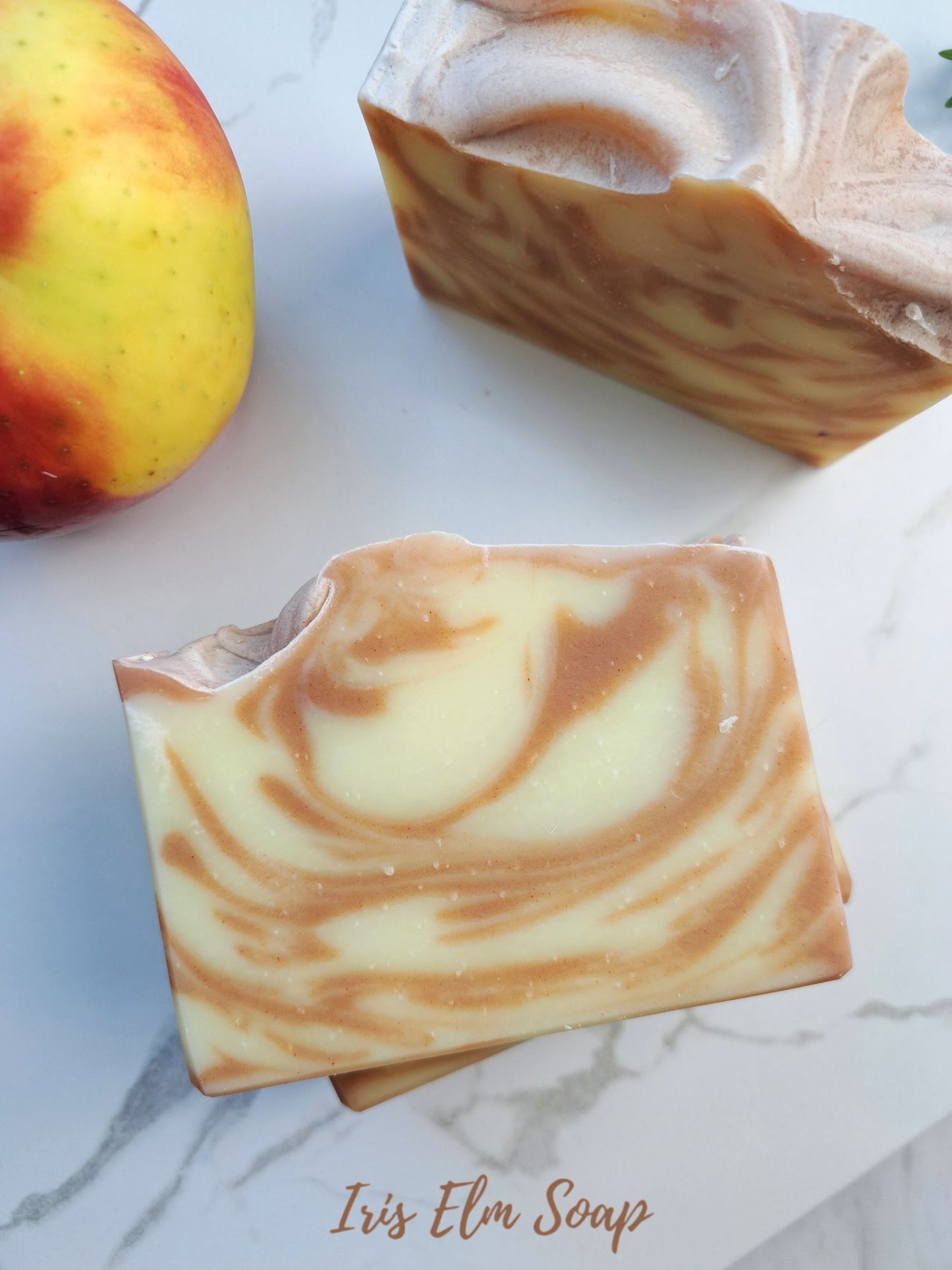 Two swirled bars of Iris Elm Soap on a light marble surface, with a red and yellow apple partially visible on the left. The soap has a cream and light brown marbled pattern.