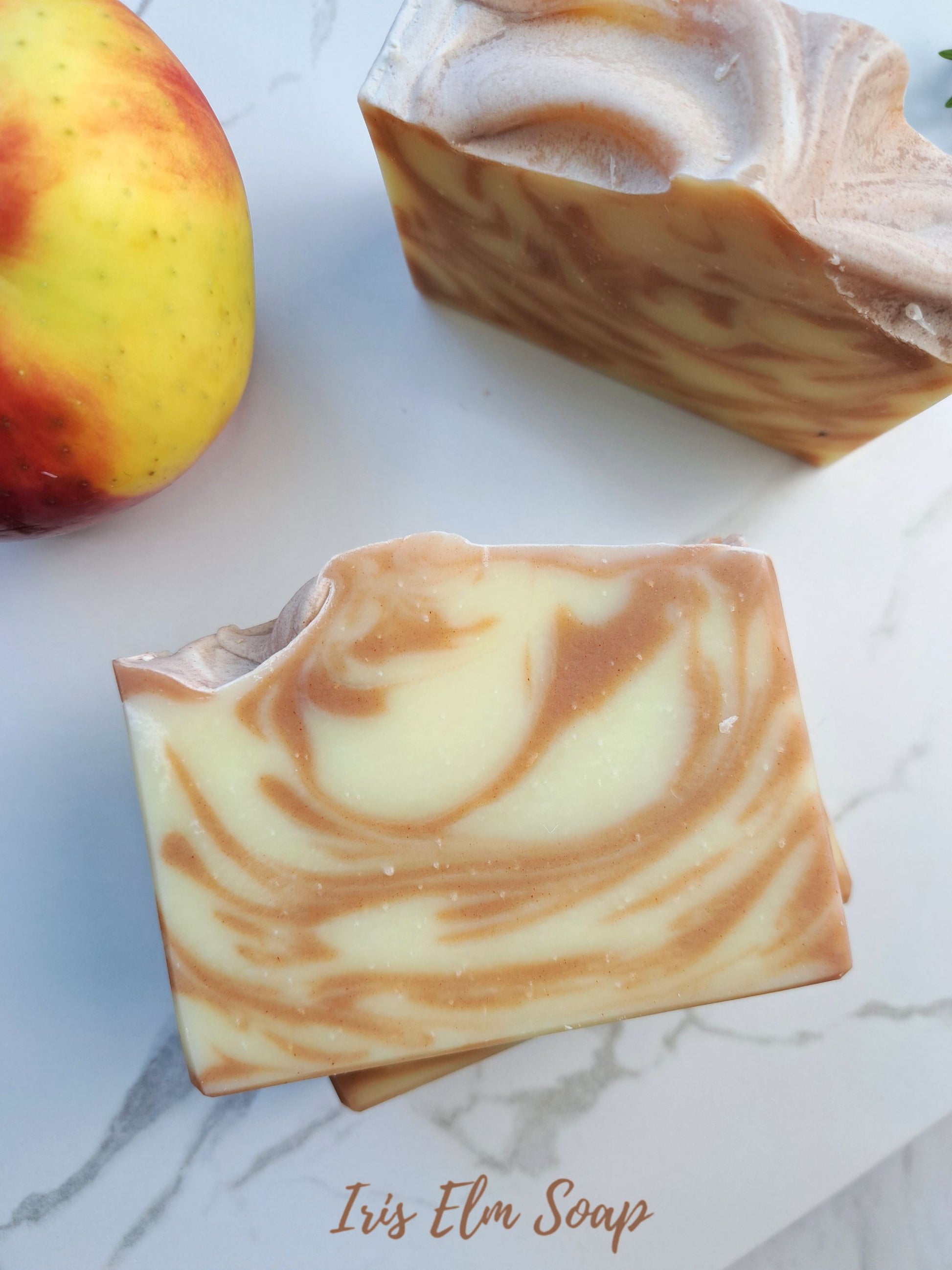 Two swirled bars of Iris Elm Soap on a light marble surface, with a red and yellow apple partially visible on the left. The soap has a cream and light brown marbled pattern.