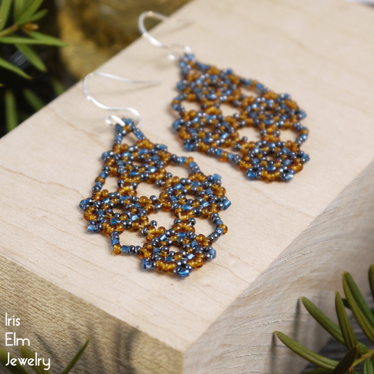 Beaded glass amber orange and blue seed bead lattice earrings on a wooden surface with greenery