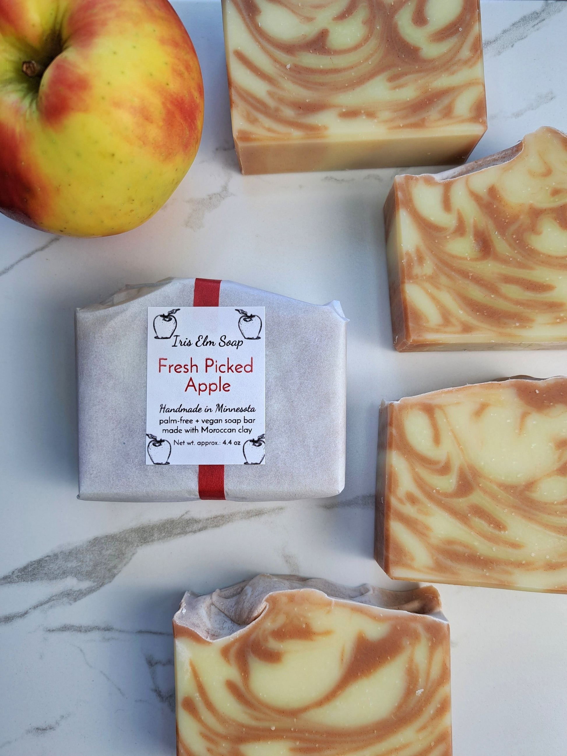 Three bars of swirled apple scented soap and one wrapped bar labeled 'Fresh Picked Apple' by Iris Elm Soap, arranged on a white marble surface next to a fresh apple.
