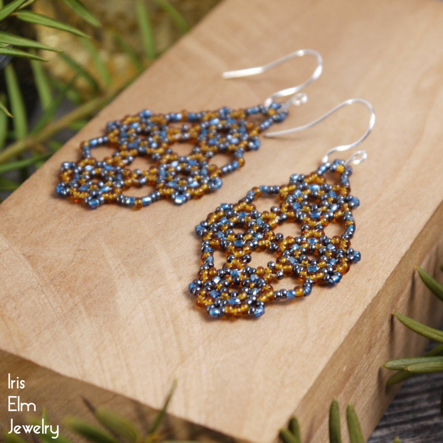 Pair of amber orange and blue seed bead lattice woven earrings on a wooden block with greenery in the background.