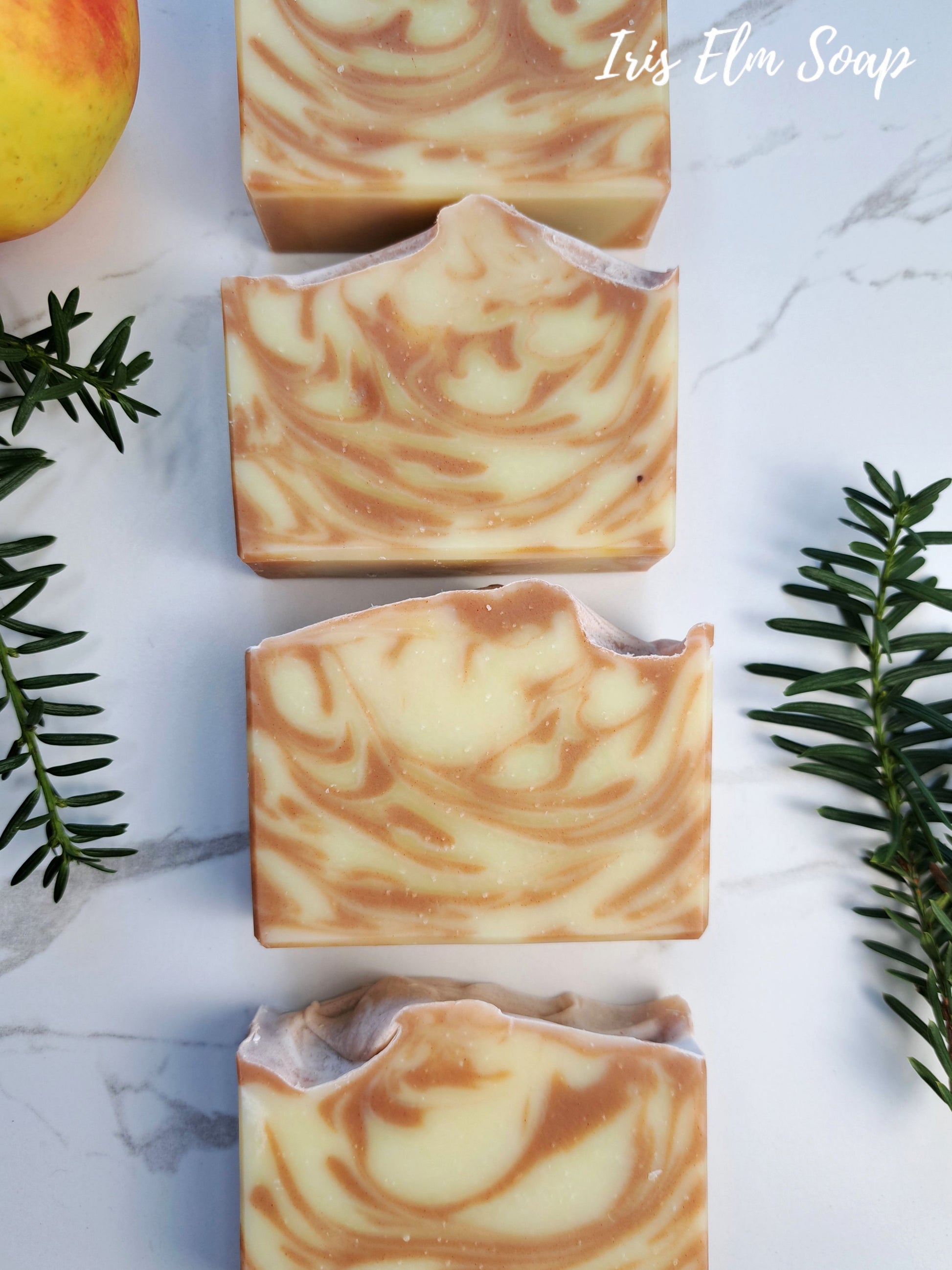 Four rectangular bars of swirled oatmeal-and-cream apple soap are arranged vertically on a white marble surface, with green sprigs and part of a peach visible as styled props. The text 'Iris Elm Soap' appears in the top right.
