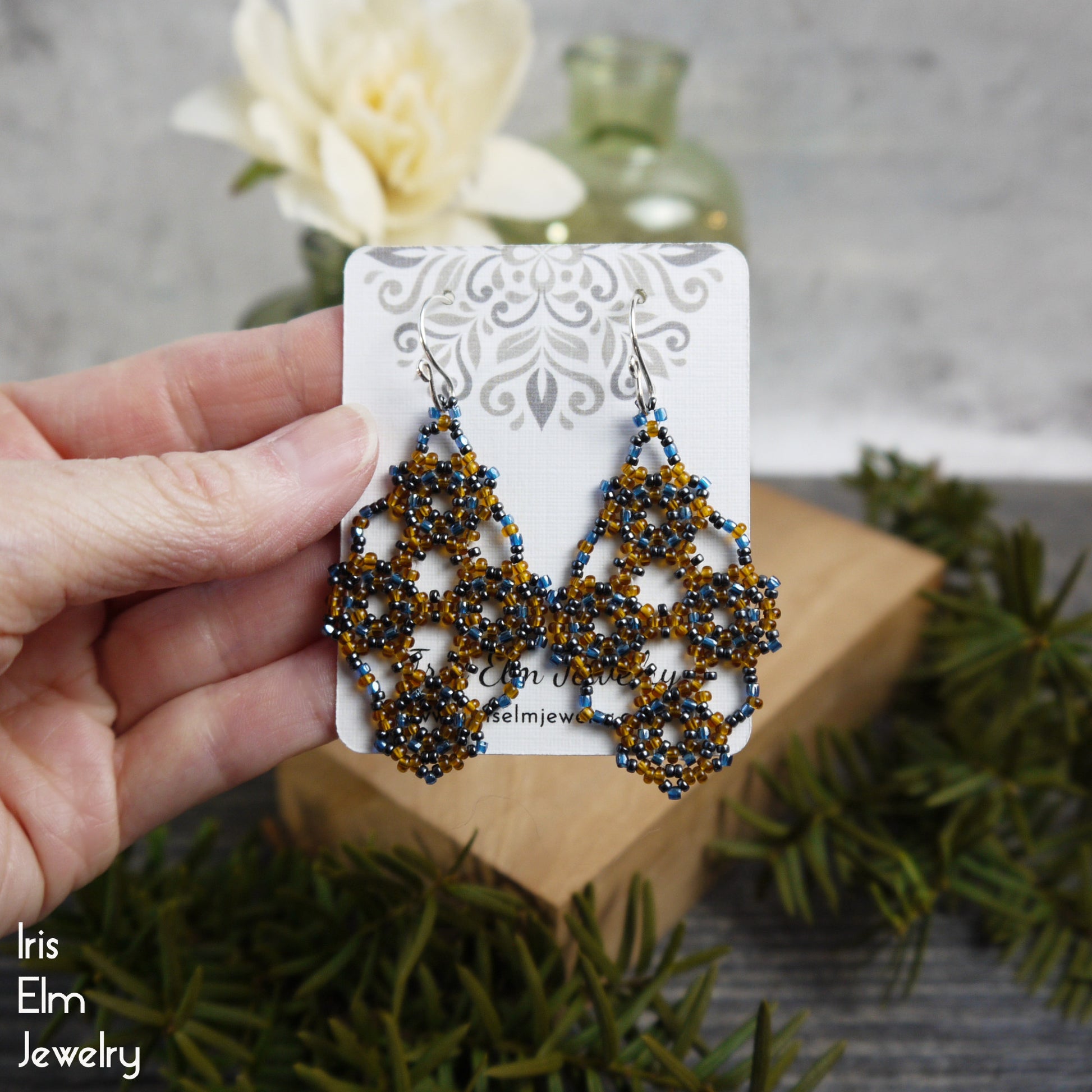 Hand holding a pair of amber orange and blue seed bead lattice beaded earrings on a branded card with a blurred background