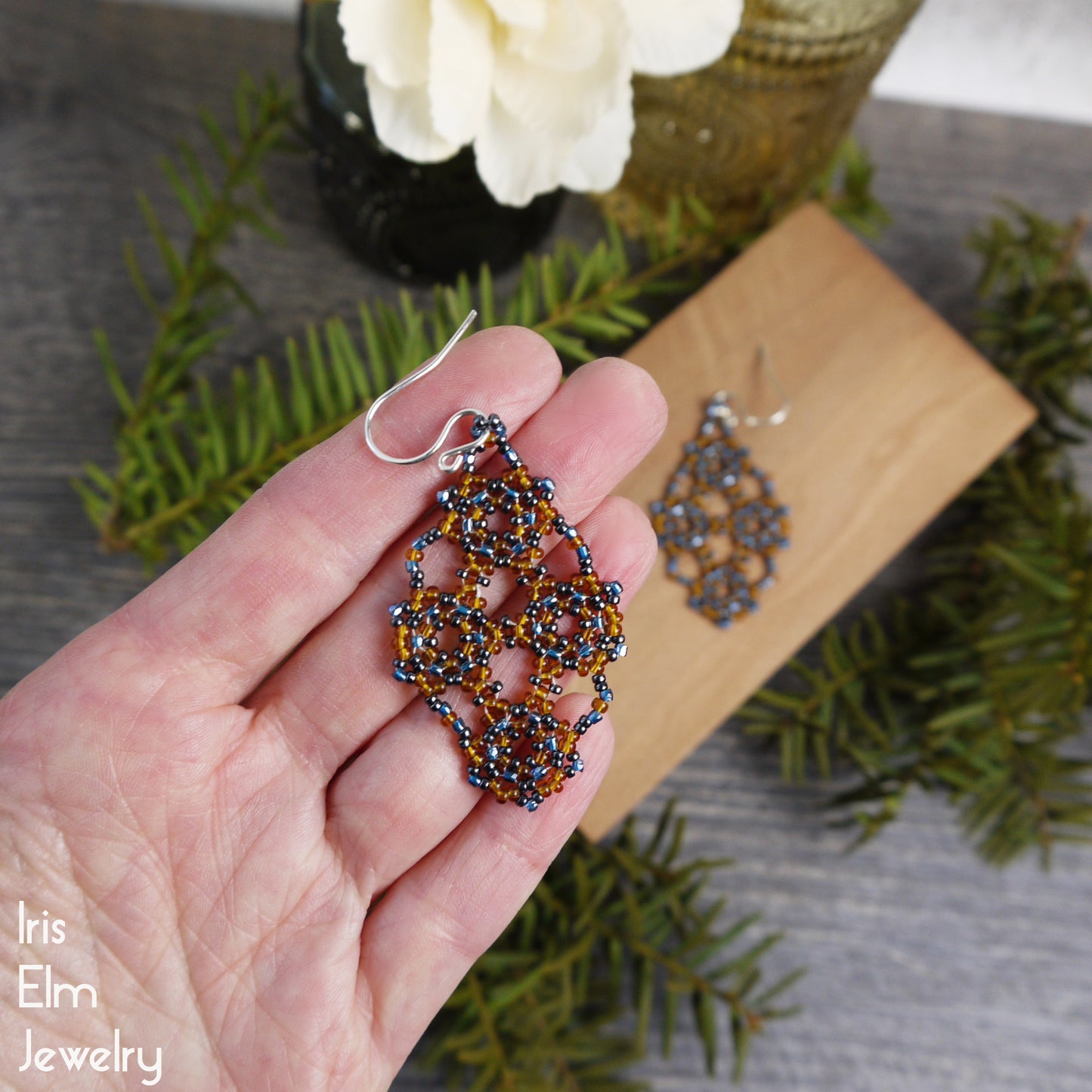 Hand holding a beadwork amber orange and blue seed bead lattice earring with sterling silver ear wires and greenery in the background, featuring 'Iris Elm Jewelry'.