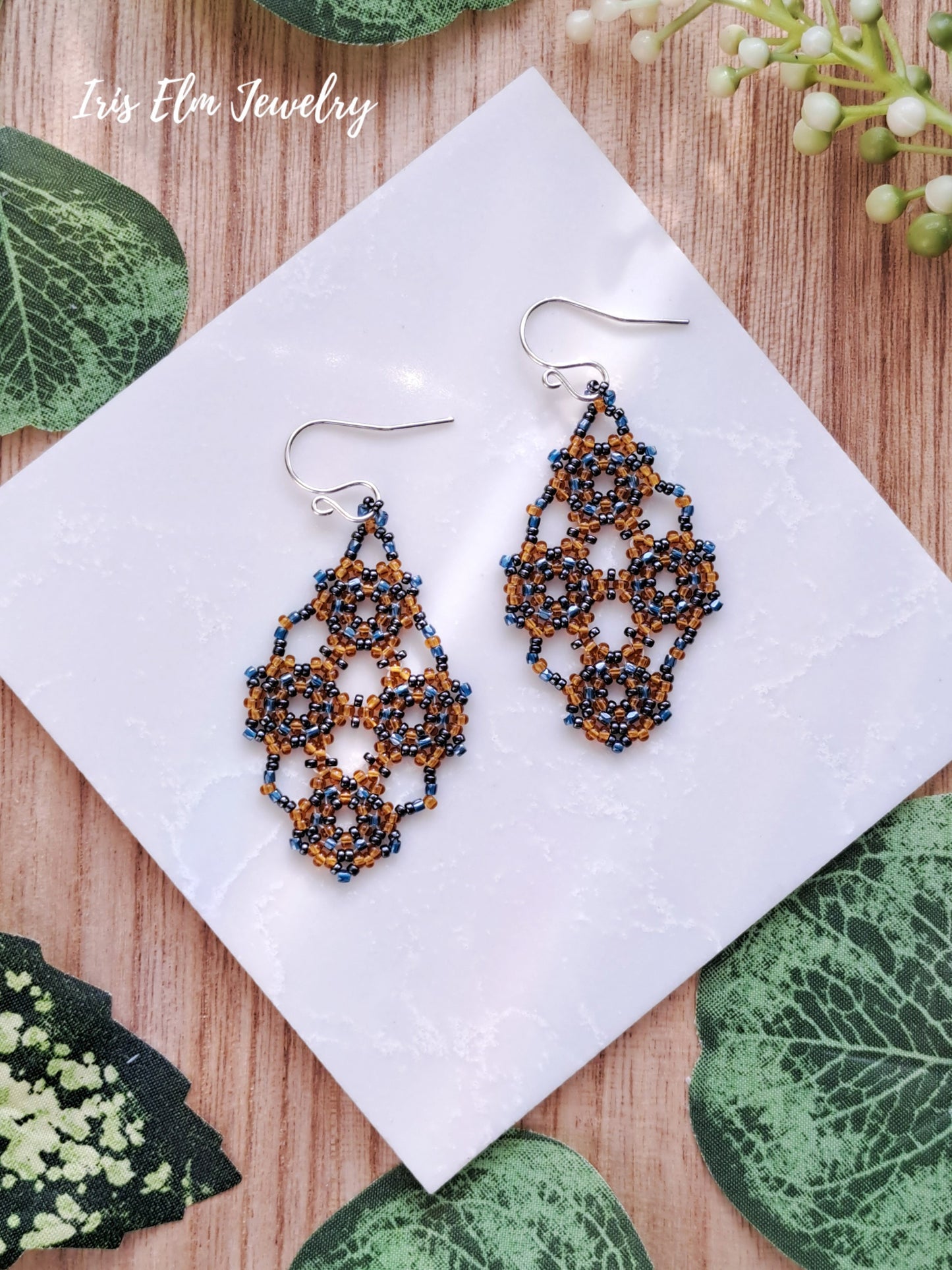 Pair of beaded orange and blue lattice seed bead earrings on a white square with green leaves around