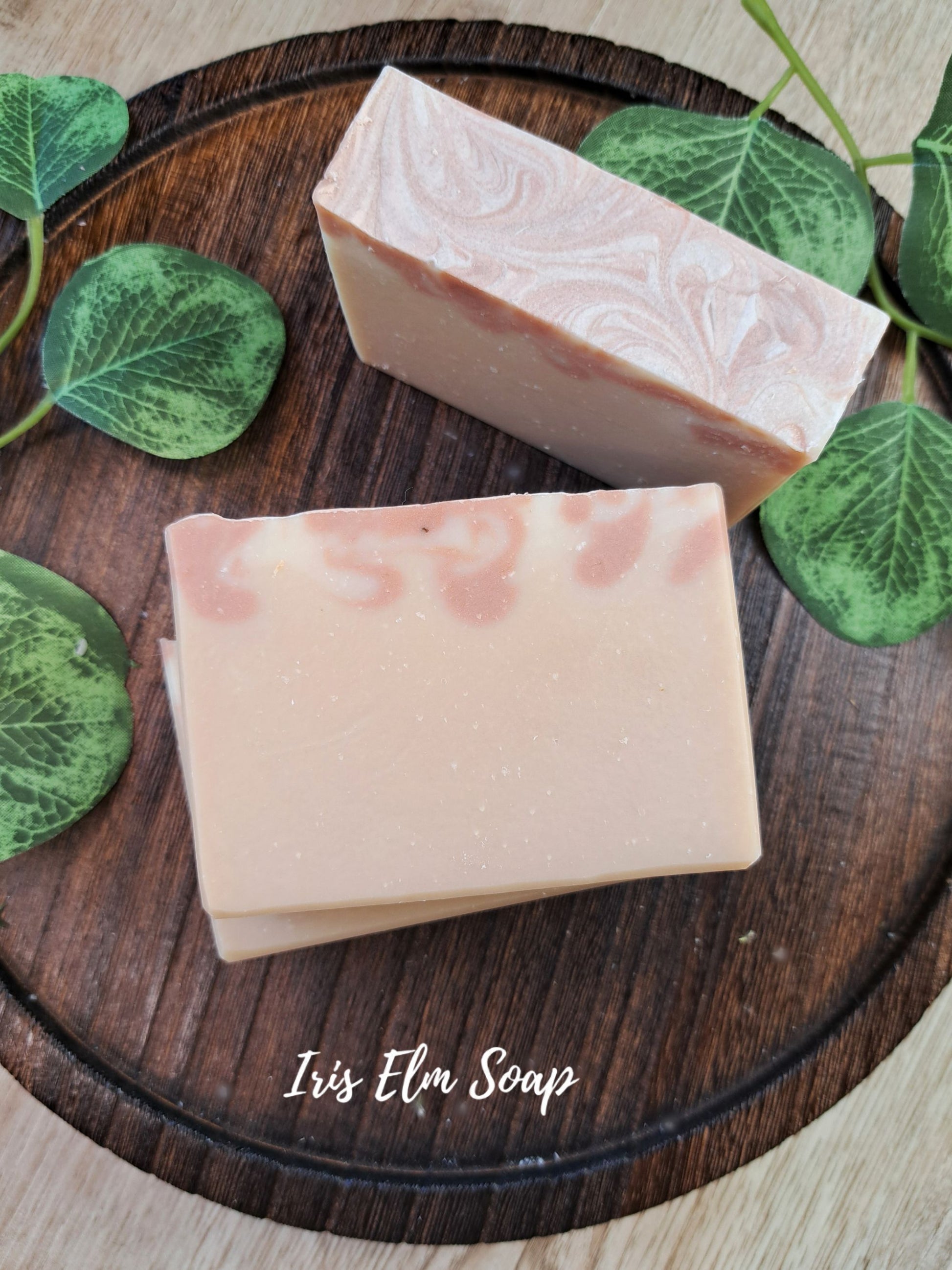 Two rectangular almond scented bars of Iris Elm Soap in a dusty pink shade with pink swirls, displayed on a dark wooden serving board decorated with green leaves. The brand name 'Iris Elm Soap' is written in white script at the bottom.