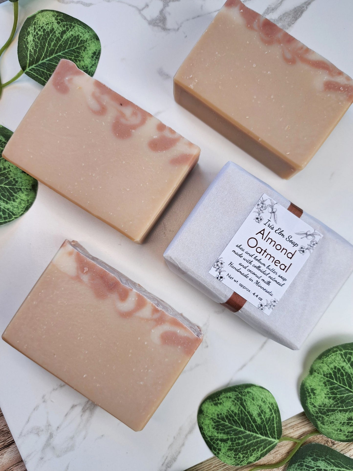 Three rectangular bars of handmade soap with a light beige base and pink swirls are arranged on a white marble surface alongside a white gift box labeled 'Almond Oatmeal'. Green leafy stems are placed around the items as styling props.