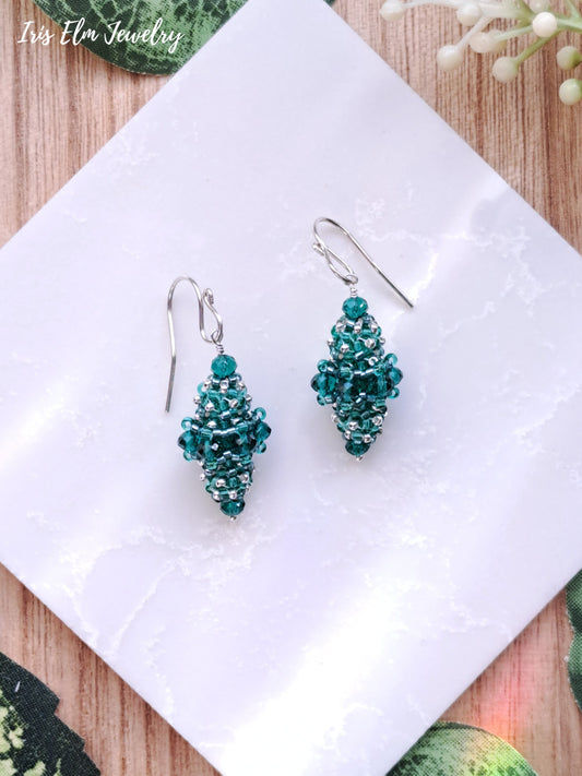 Pair of teal green glass beaded bead earrings on a white cloth with 'Iris Elm Jewelry' branding.