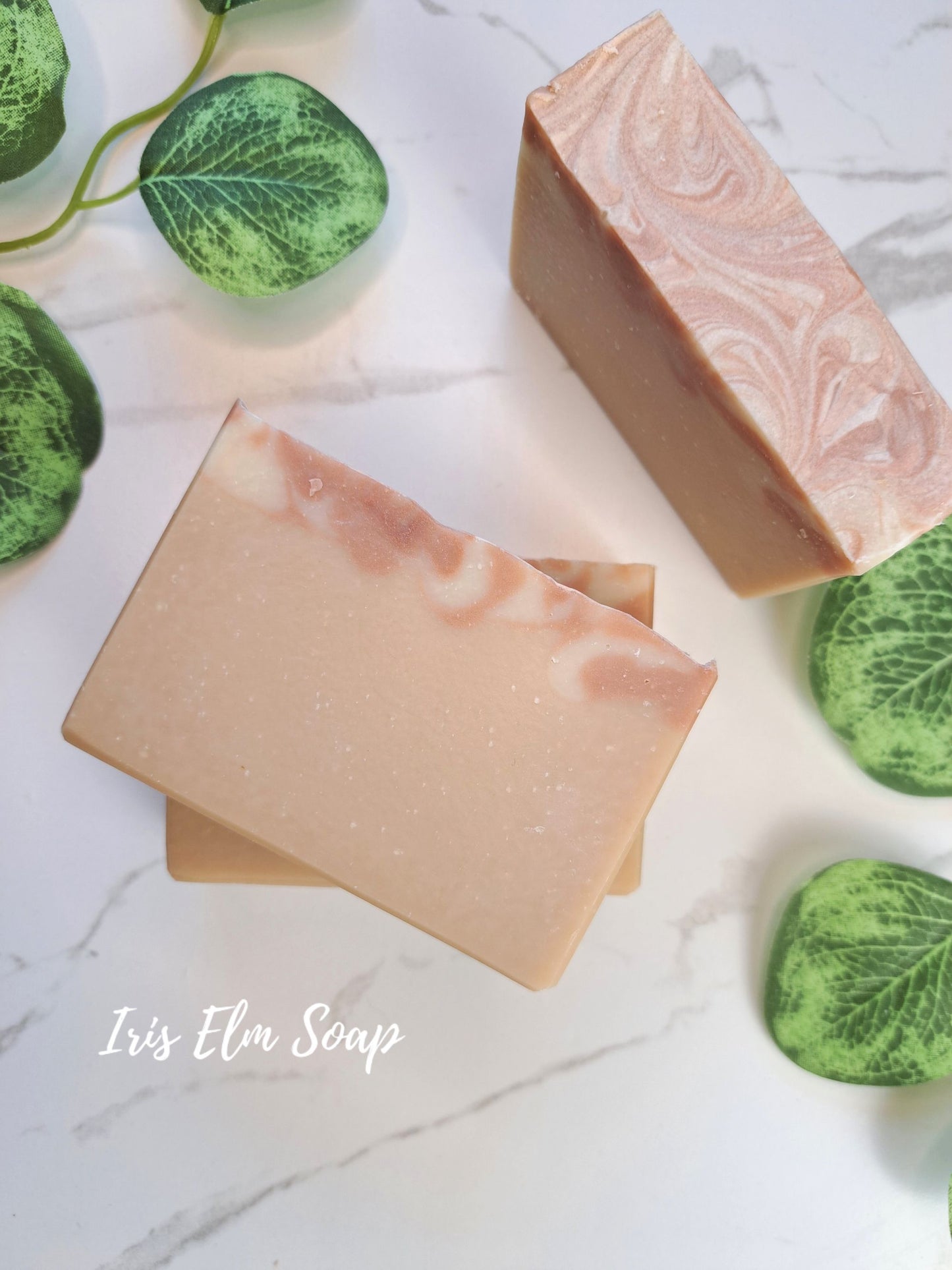 Two rectangular almond scented bars of Iris Elm Soap with a light peach and cream marbled swirl pattern, styled on a white marble background with green leaves.