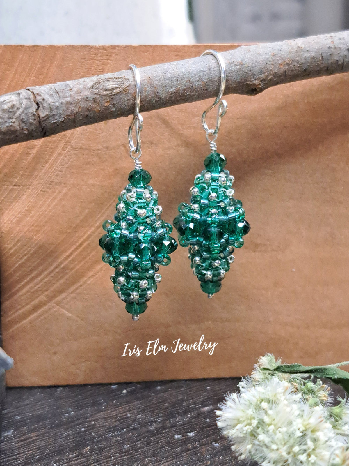 A pair of beaded dangle earrings in shades of green and silver, hanging from a rustic branch. The background is a textured brown paper sheet with a wooden surface below, and a small white flower is in the bottom right corner.
