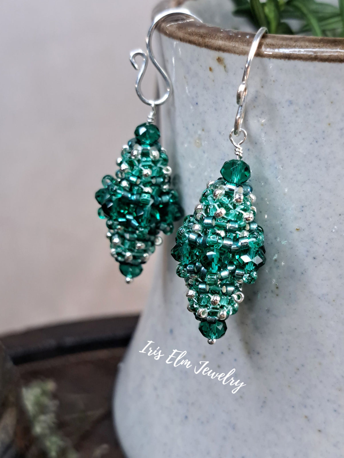 A pair of handmade beaded dangle earrings in emerald green with silver metal findings, hanging from a speckled gray ceramic pot.