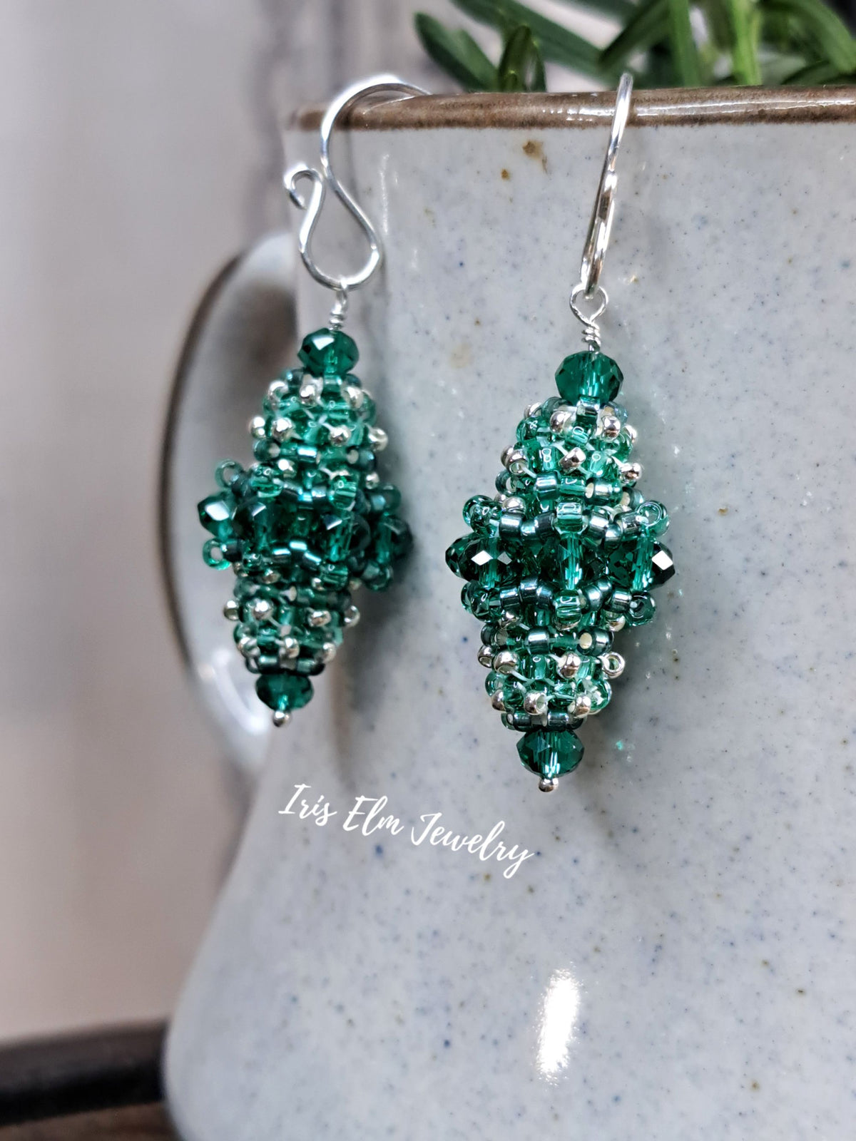 A pair of handmade beaded dangle earrings in shades of emerald and teal green with silver-toned metal hooks, displayed against a speckled ceramic mug with a 'Iris Elm Jewelry' watermark.
