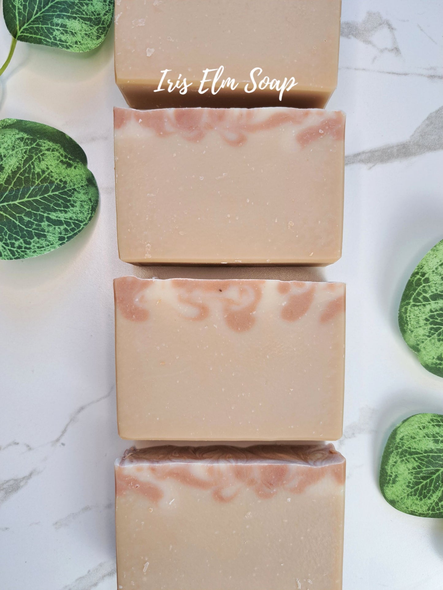 Four rectangular bars of Iris Elm Soap arranged vertically on a white marble surface, decorated with green leaves. The soaps are a light peach-beige color with pinkish swirls near the top.