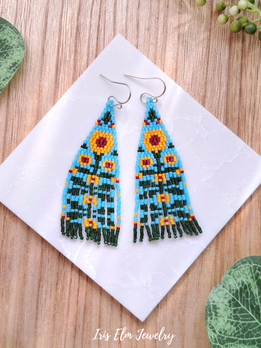 beadwoven yellow sunflower with green and light blue beads floral folk art seed bead fringe earrings with greenery background and Iris Elm Jewelry branding