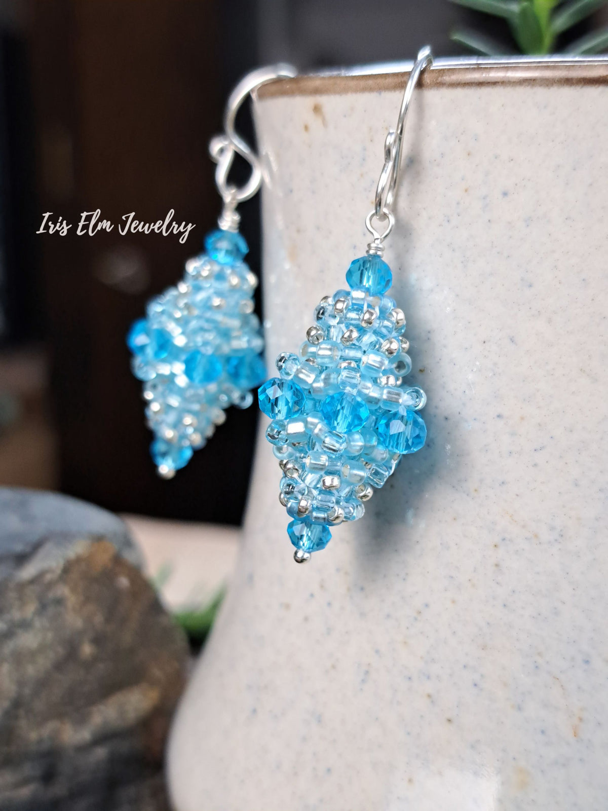 A pair of handmade dangle earrings by Iris Elm Jewelry, featuring light blue faceted beads and silver-toned metal hooks, hanging against a textured ceramic vase.