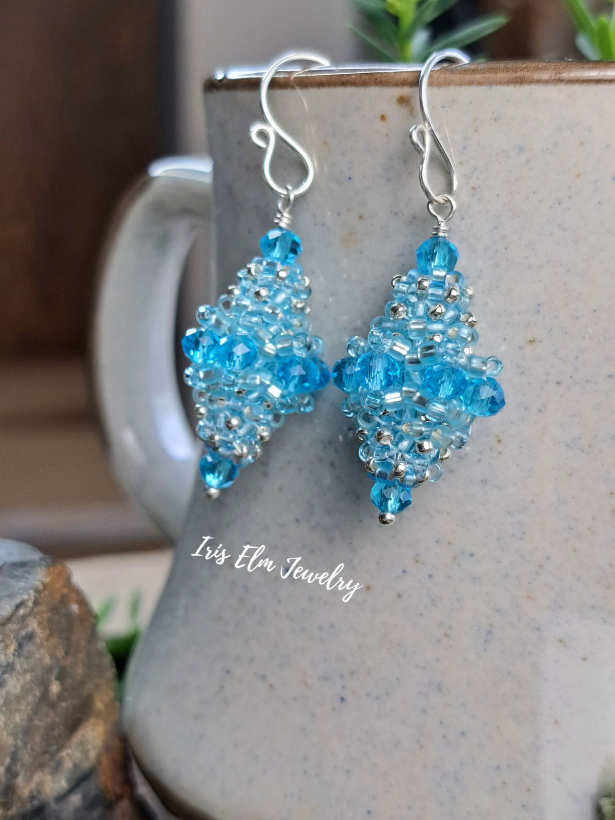 A pair of handmade beaded dangle earrings with light blue faceted beads and silver hooks, hanging in front of a speckled ceramic mug. The text 'Iris Elm Jewelry' is visible on the mug.