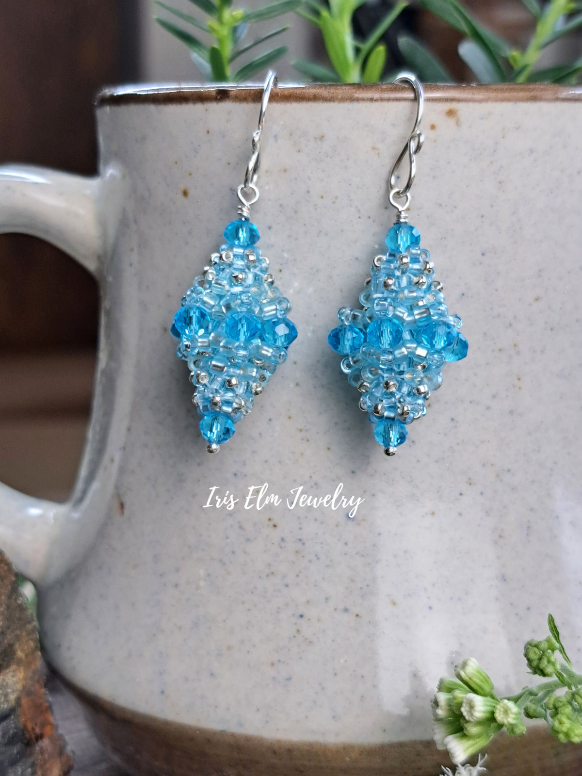 A pair of handmade beaded dangle earrings in bright blue and clear beads with silver hooks, displayed on a speckled ceramic mug with green foliage in the background. The text 'Iris Elm Jewelry' is visible as a watermark.