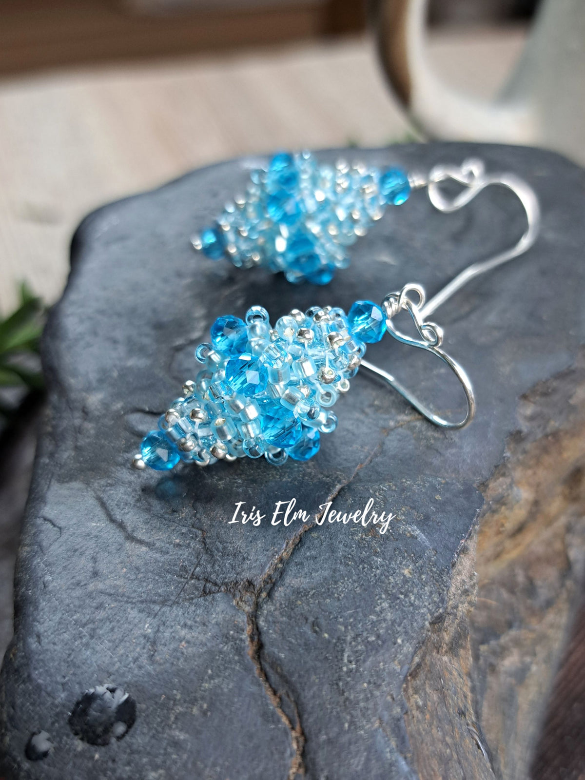 A pair of handmade beaded earrings with light blue faceted beads and silver hooks, displayed on a dark textured stone surface. The brand name 'Iris Elm Jewelry' is watermarked on the stone.