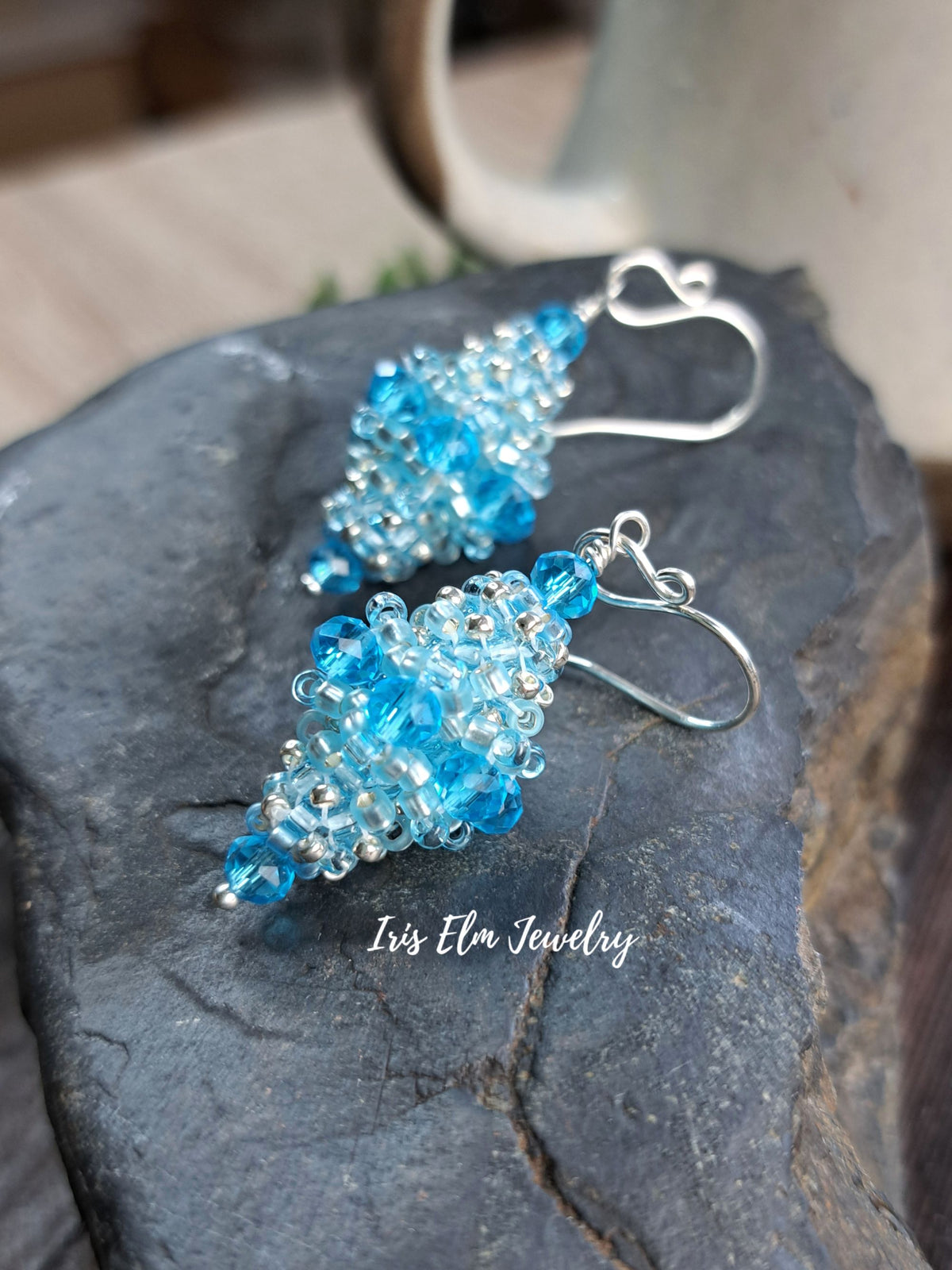 A pair of handmade beaded dangle earrings with light blue and clear faceted beads on silver fishhook wires, displayed on a dark slate stone with a ceramic mug in the blurred background. The text 'Iris Elm Jewelry' is overlaid on the image.
