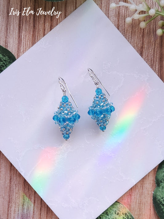 light Blue beaded bead glass earrings on a white card with a rainbow effect, on a wooden surface.