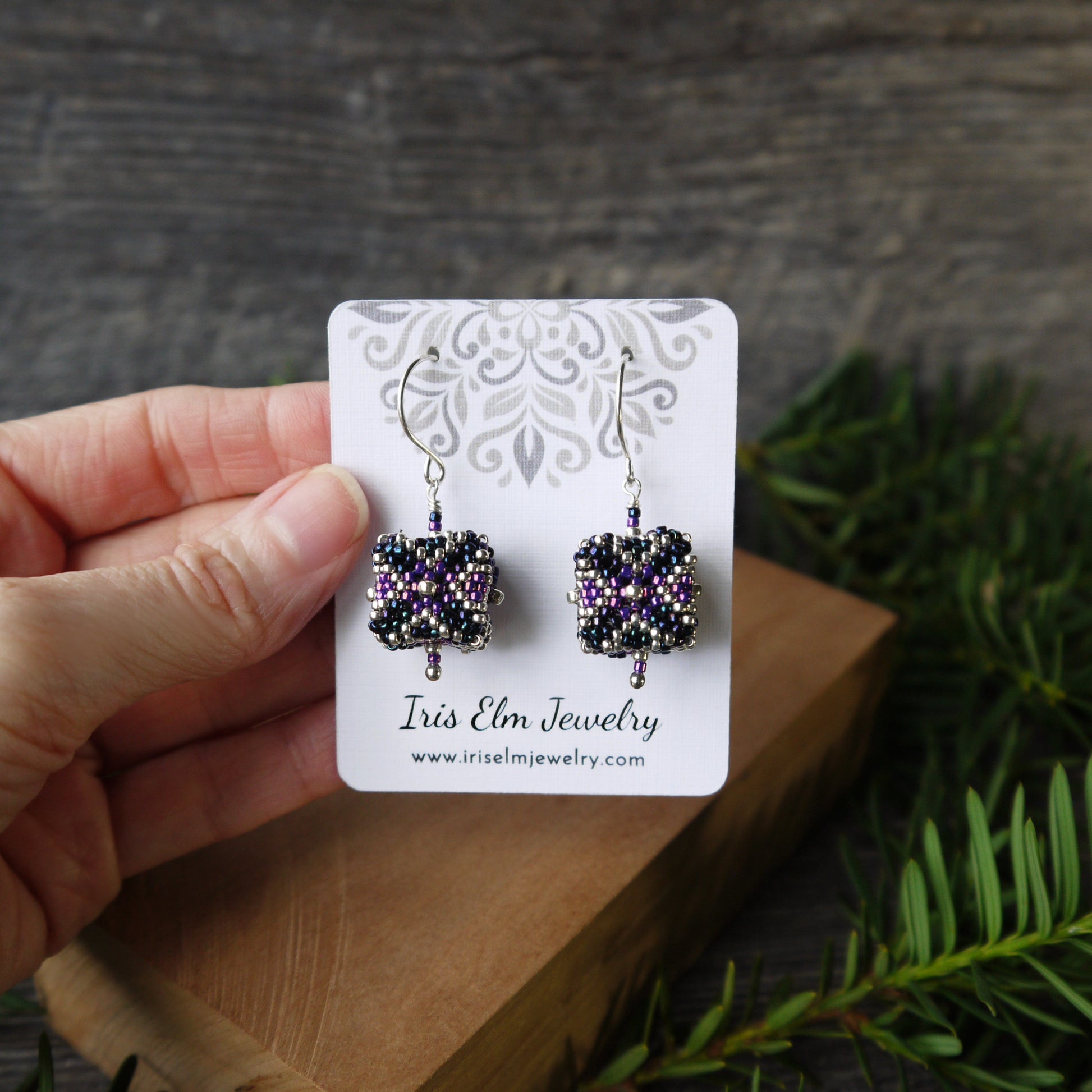 Hand holding a card with cube shaped seed bead beaded earrings on a wooden surface with greenery