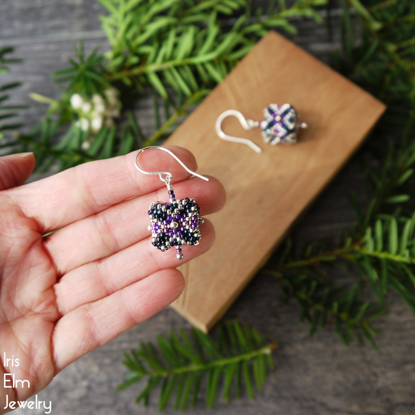 Hand holding a pair of beaded purple and blue cube seed bead beadwork earrings with a wooden block and greenery in the background.