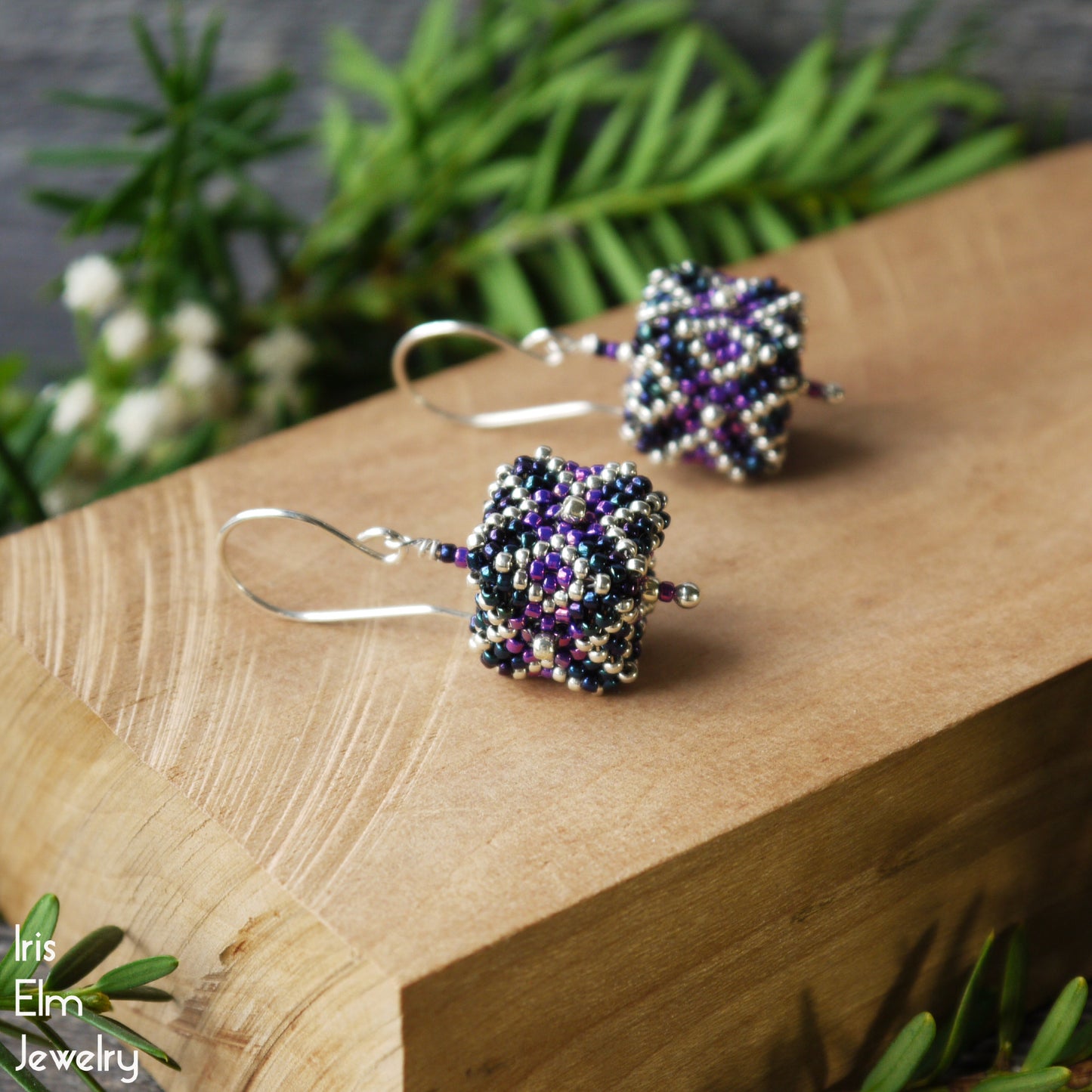 Beaded purple and blue seed bead cube shaped earrings with sterling silver ear wires on a wooden surface with greenery in the background