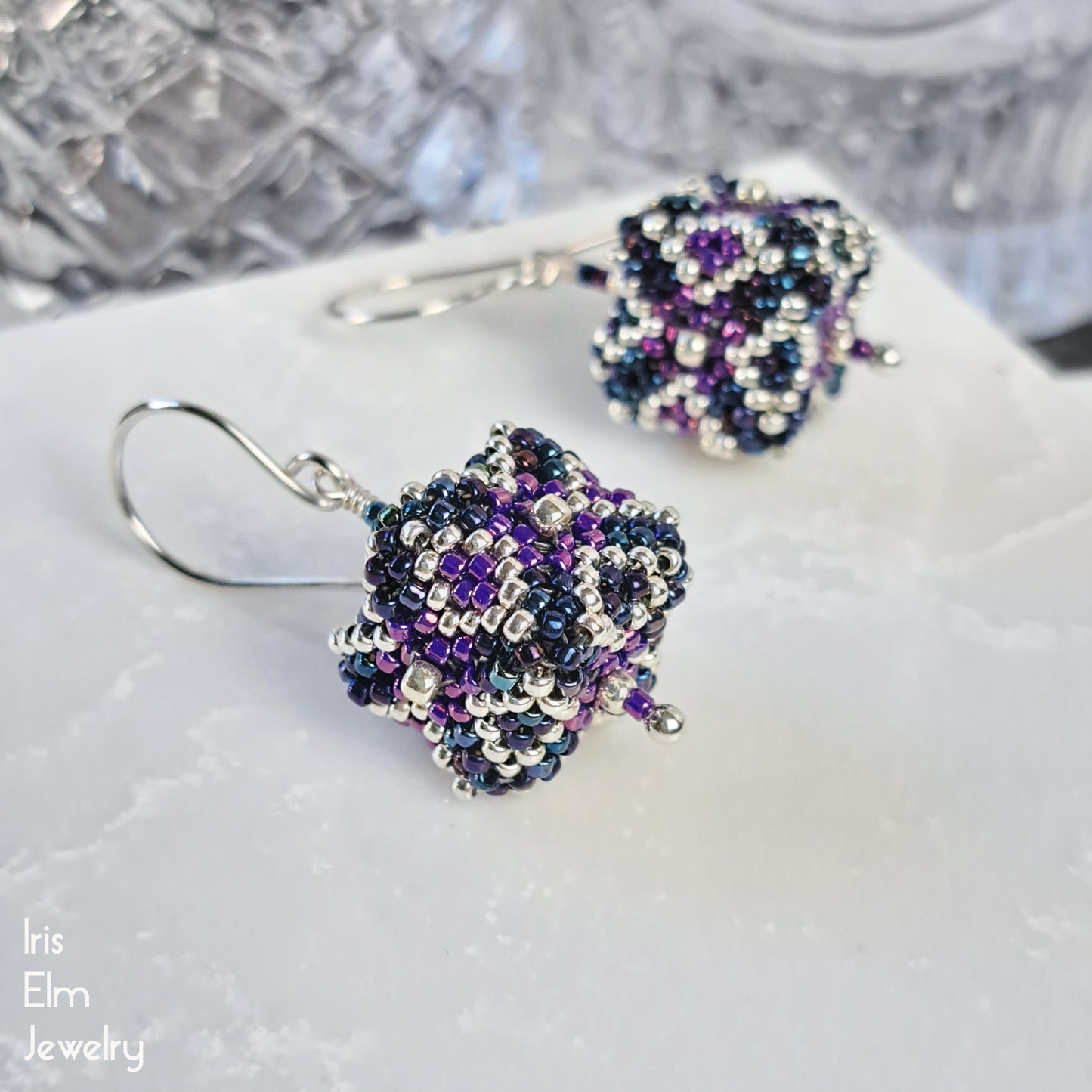 iridescent purple, dark blue, and silver glass seed bead 3d cube earrings on a white surface with 'Iris Elm Jewelry' branding.