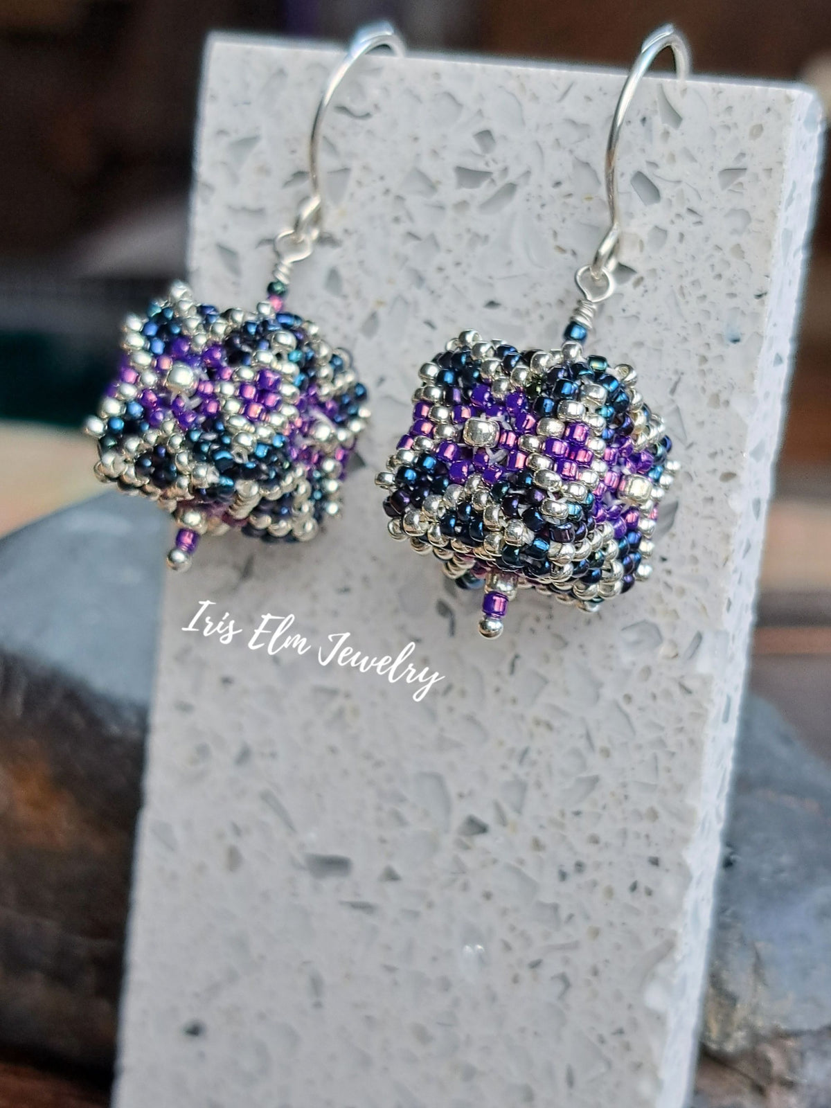 A pair of handmade beaded drop earrings with silver fishhook wires, featuring a textured cube design in purple, iridescent blue, and silver seed beads. The earrings are displayed on a light gray stone block with a watermark reading 'Iris Elm Jewelry' on the side.