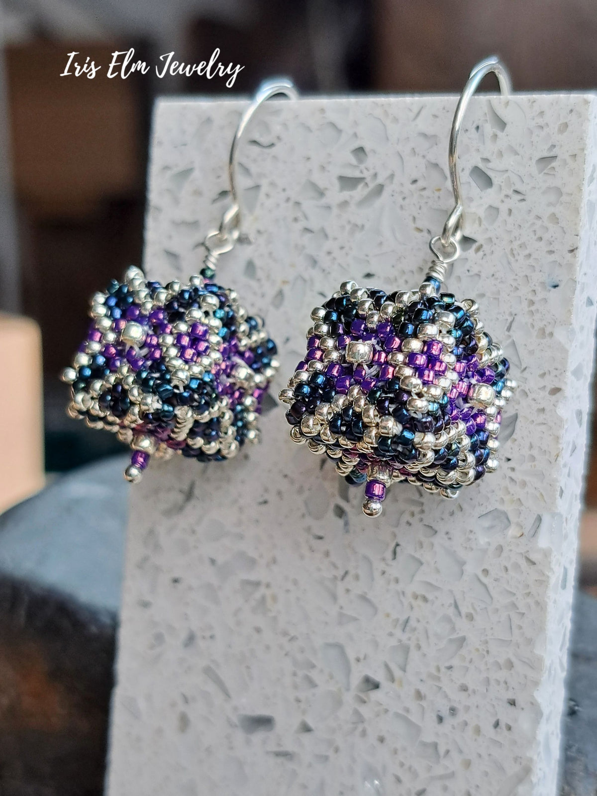 A pair of cube beaded dangle earrings with silver fishhook wires, featuring intricate patterns of purple, iridescent blue, and silver seed beads. The earrings are displayed on a textured white stone block with a 'Iris Elm Jewelry' logo overlay in the top left corner.