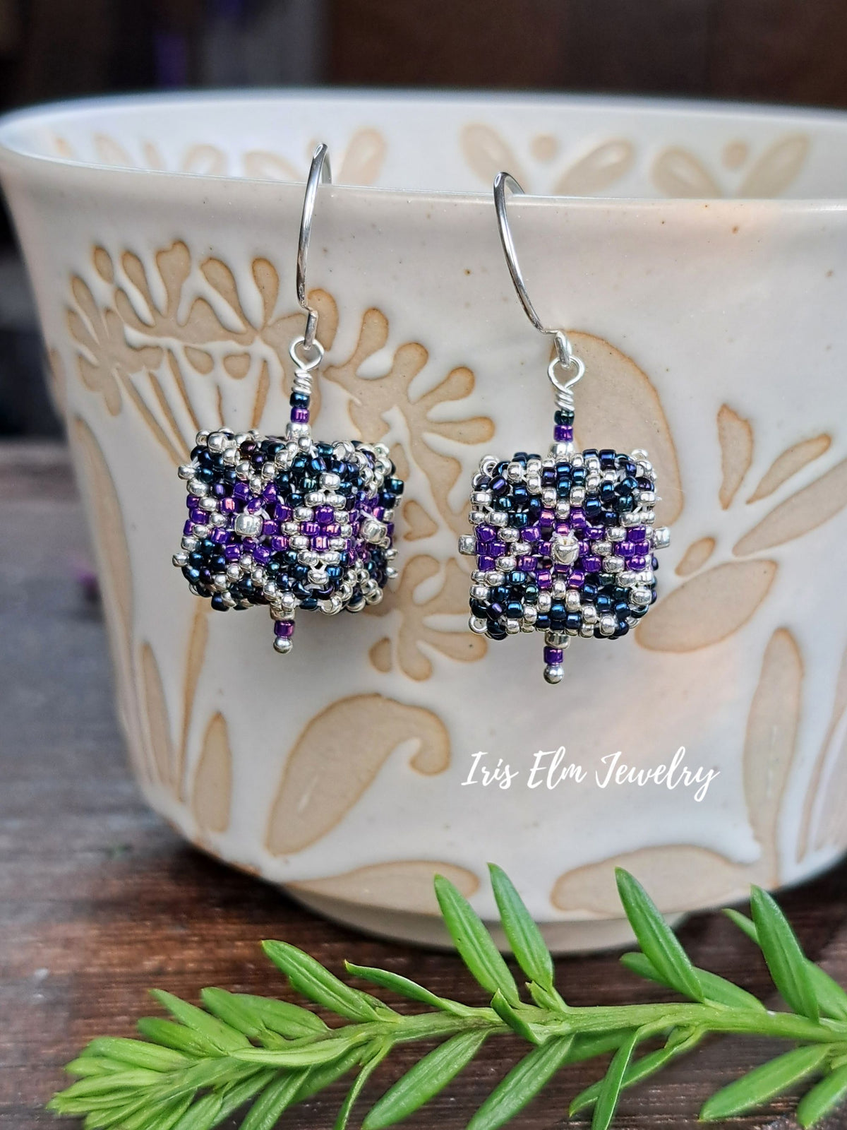 Square beaded drop earrings with purple, navy, and silver beads, hanging in front of a white ceramic bowl with beige floral patterns. A green sprig is in the foreground, and the text 'Iris Elm Jewelry' is visible on the bowl.