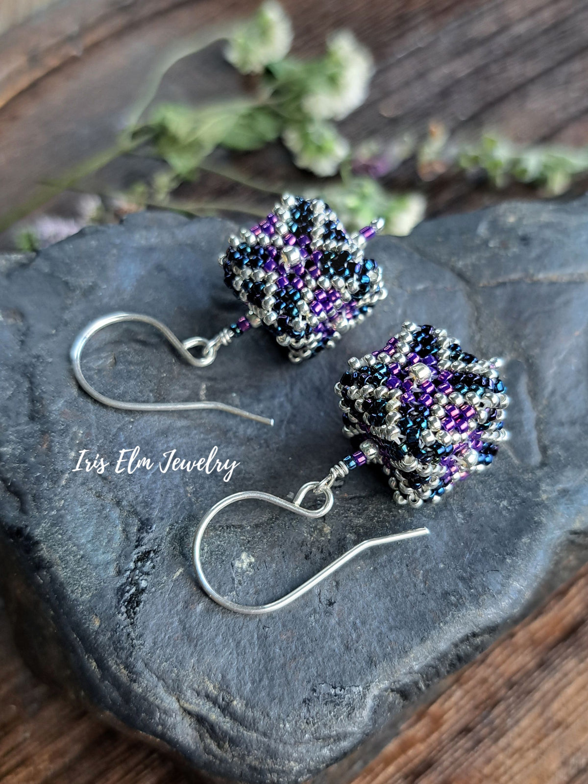 A pair of handmade beaded dangle earrings with silver fishhook wires, displayed on a dark slate stone. The beads are woven in silver, purple, and black, with dried flowers in the background on a rustic wooden surface.