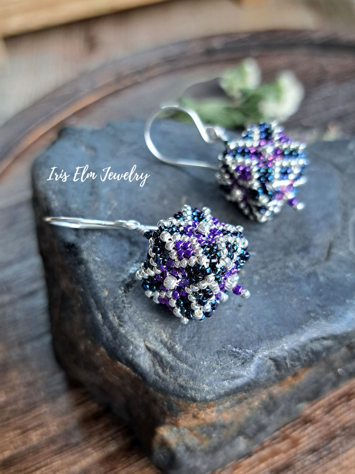Handmade beaded dangle earrings with silver fishhook wires, featuring intricate clusters of purple, black, and white seed beads. The earrings are displayed on a dark textured stone slab over a rustic wooden surface, with a watermark reading 'Iris Elm Jewelry' on the left.